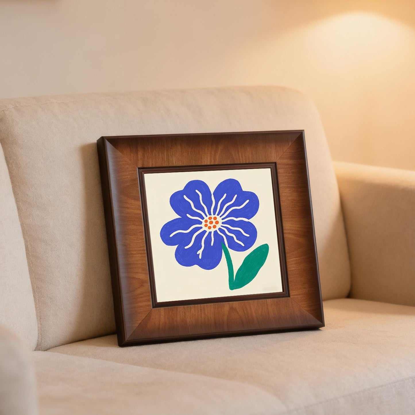 Framed blue flower illustration leaning against the back of a beige sofa in a cozy, warm-lit living room setting.