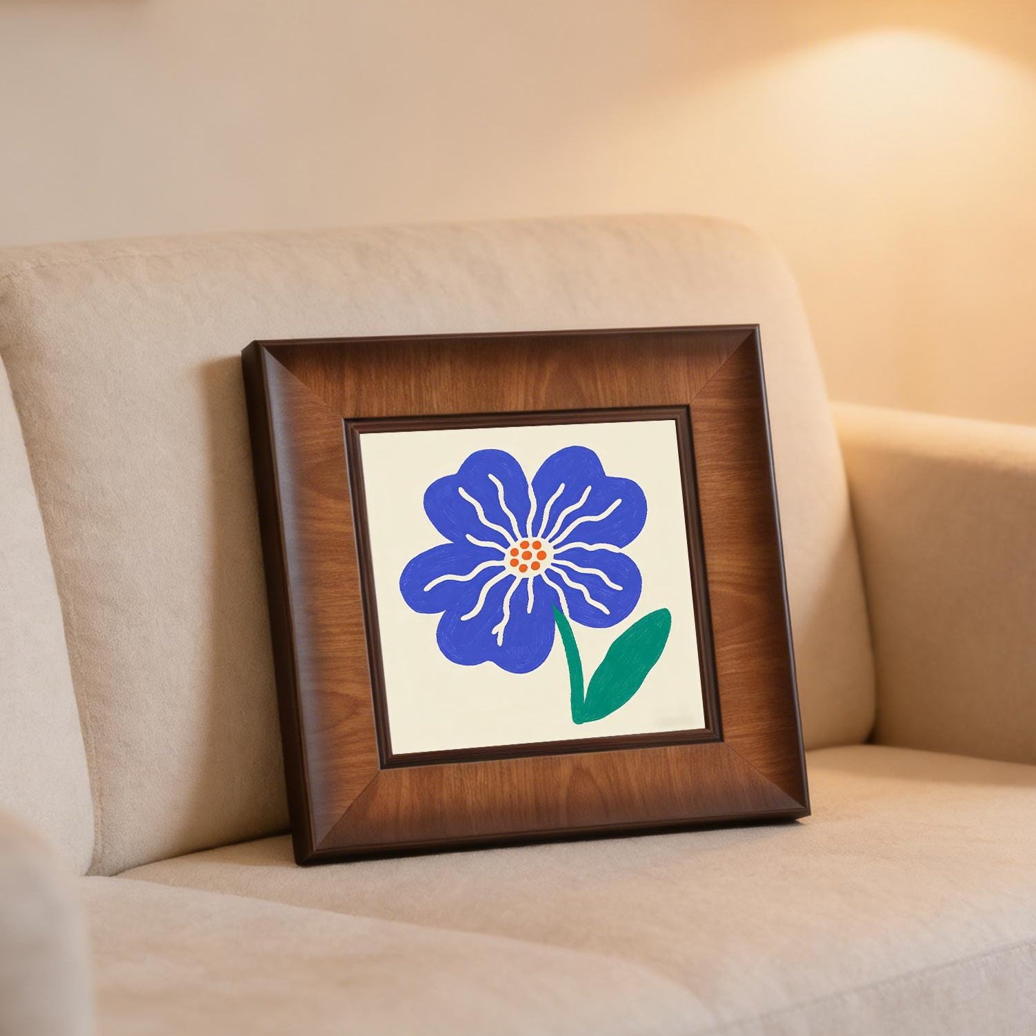 Framed blue flower illustration leaning against the back of a beige sofa in a cozy, warm-lit living room setting.