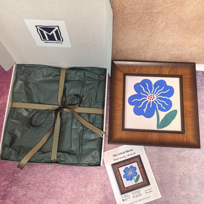 A professional unboxing set for 'The Cobalt Bloom' framed art by Miracle Walls. Features a vibrant blue flower print in a dark wood frame, a luxury white gift box with green tissue and gold ribbon, and an artist's signature certificate.