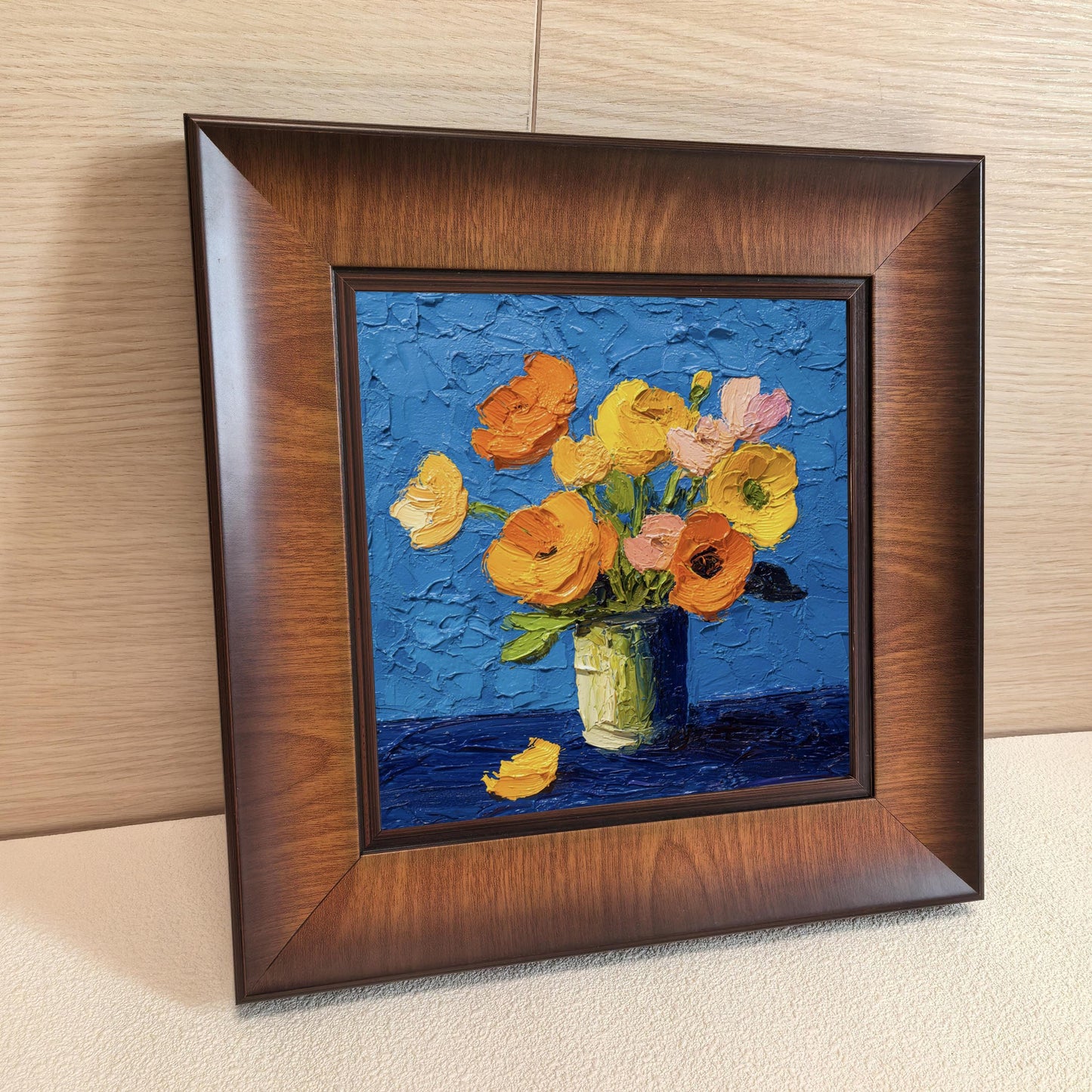 Close-up side angle of a square wooden frame containing a colorful floral oil painting, leaning against a light wood-paneled wall.