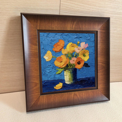 Close-up side angle of a square wooden frame containing a colorful floral oil painting, leaning against a light wood-paneled wall.