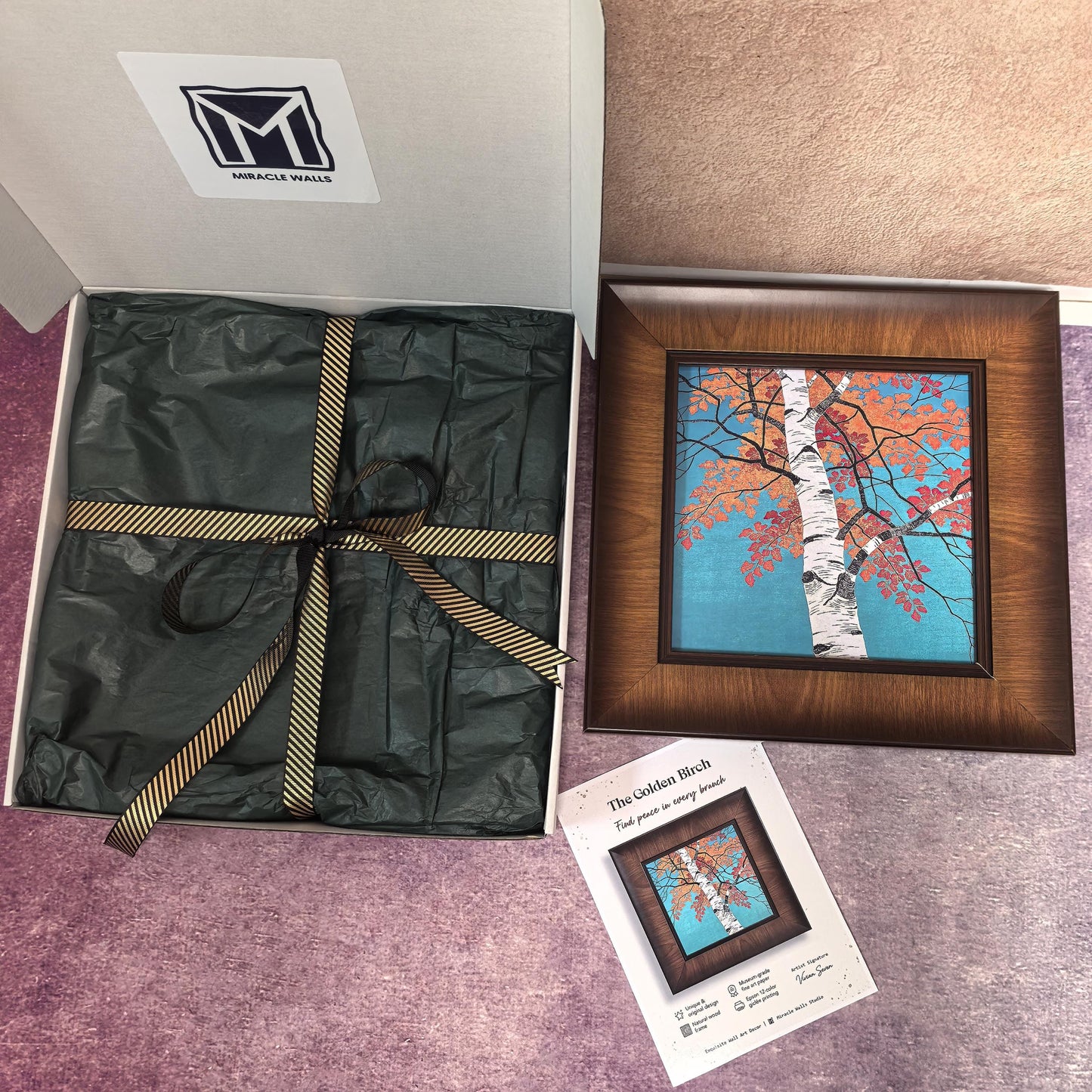 An open white gift box from Miracle Walls featuring a wrapped package in dark green tissue paper with a gold-striped ribbon. Beside it sits a framed art print titled "The Golden Birch," showing a white birch tree with orange leaves against a turquoise sky in a wide wooden frame.