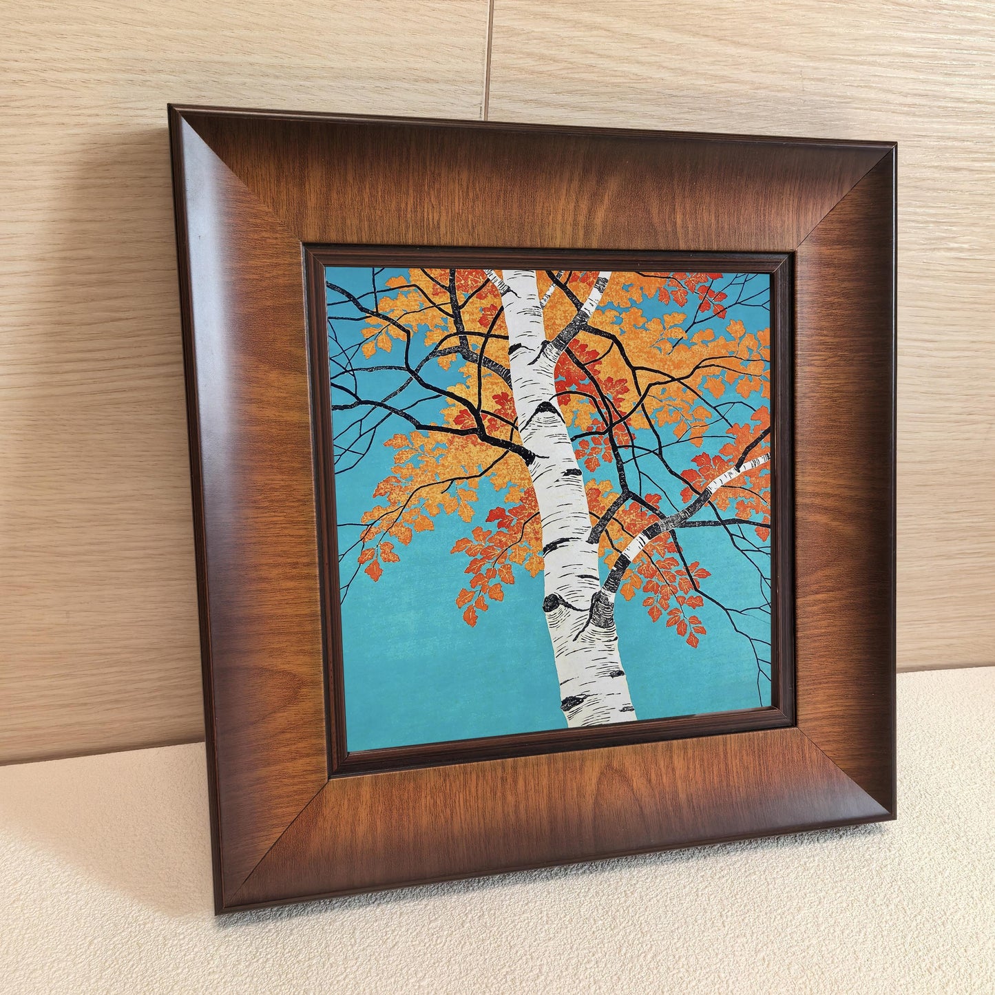 A square autumn tree art print in a dark wood frame leaning against a light-colored wooden wall, highlighting the natural texture and beveled edge of the frame.