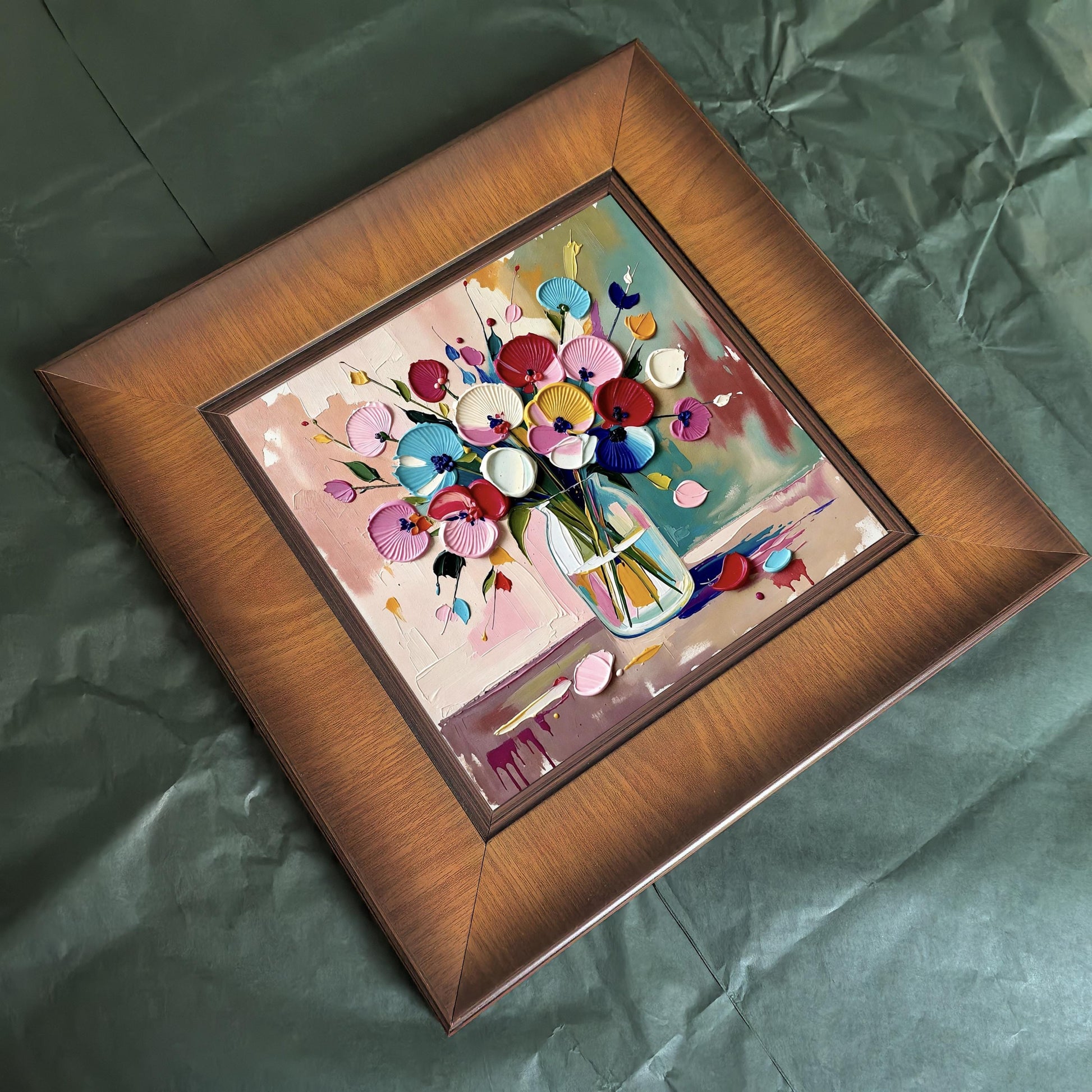Overhead view of a square wooden framed floral painting resting on a dark green textured background, showing the deep grain of the timber frame.