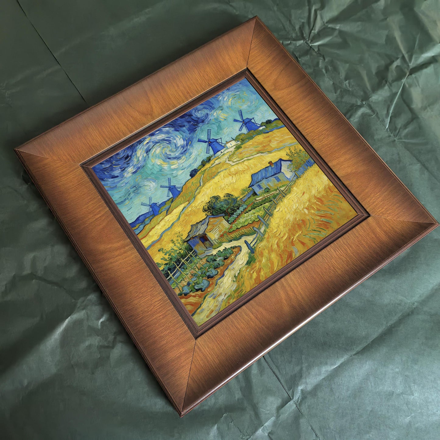 Bird's-eye view of a square wood-framed art print resting on textured dark green packaging paper, highlighting the frame's grain and mitered corners.