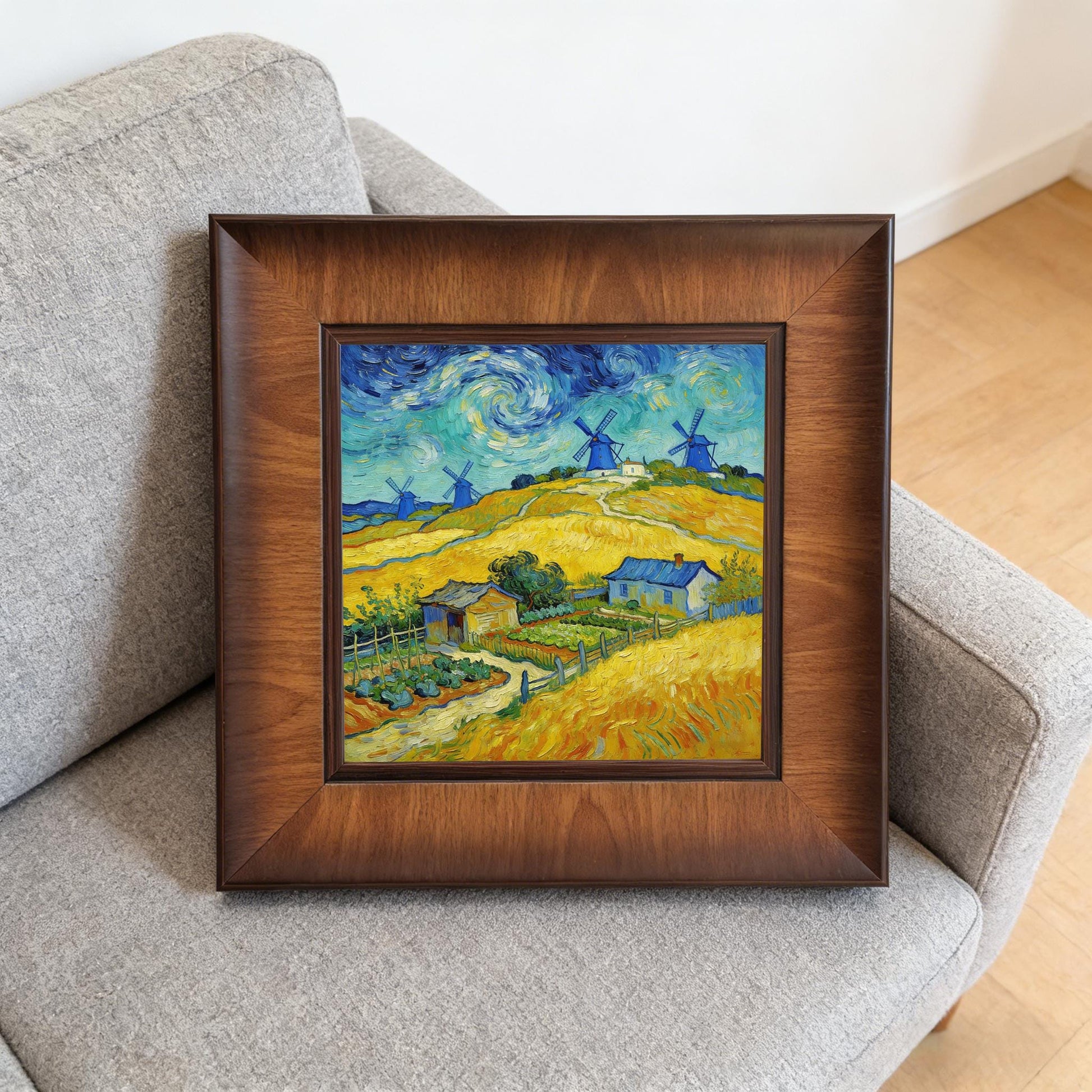A square wood-framed painting of blue windmills and yellow fields resting on a grey fabric sofa, demonstrating how the art looks in a casual living space.