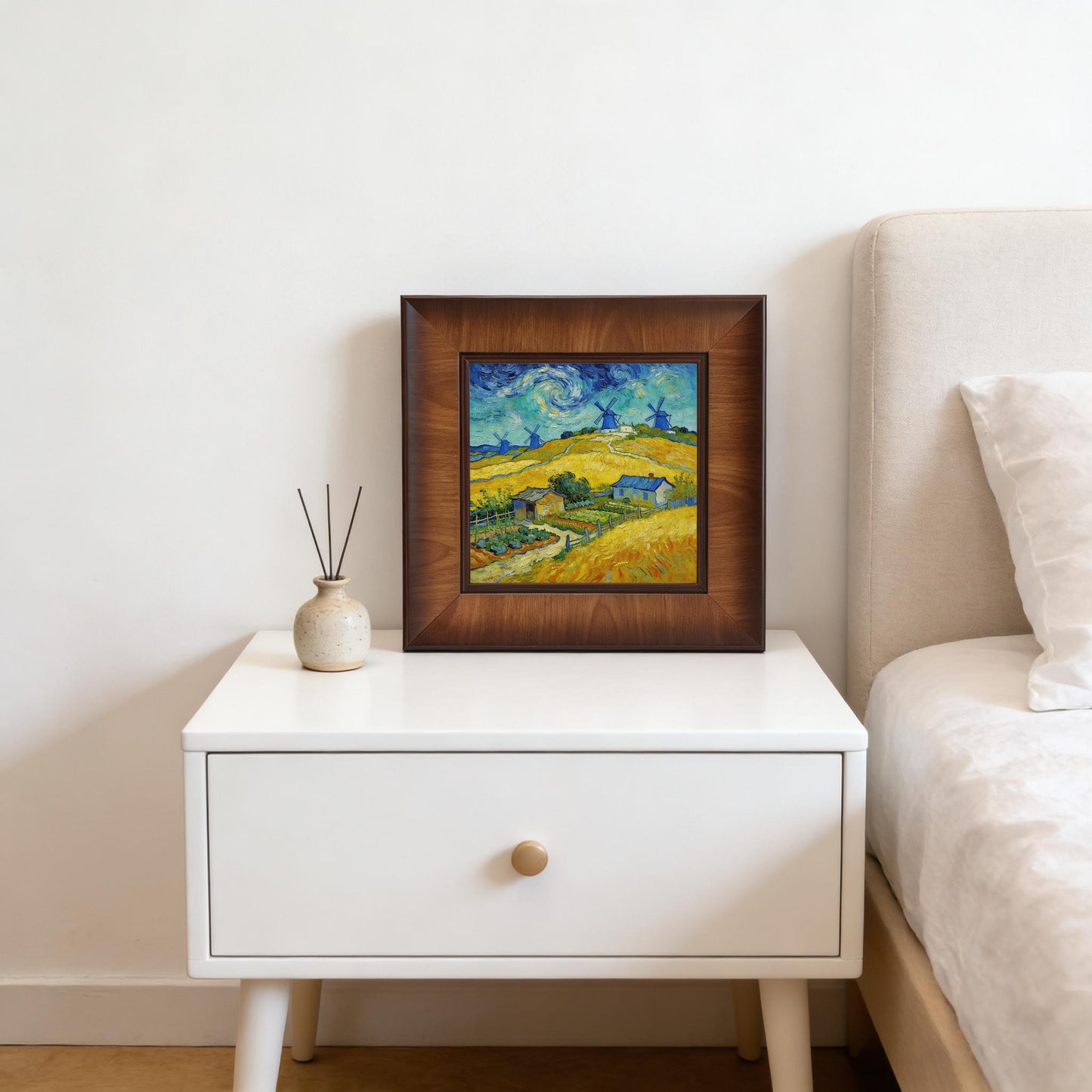 Square framed landscape art leaning on a white mid-century modern nightstand next to a bed, styled with a small ceramic vase and scent diffusers.