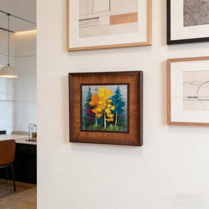 A square wooden framed painting of colorful trees displayed as part of a modern gallery wall in a bright, minimalist interior.