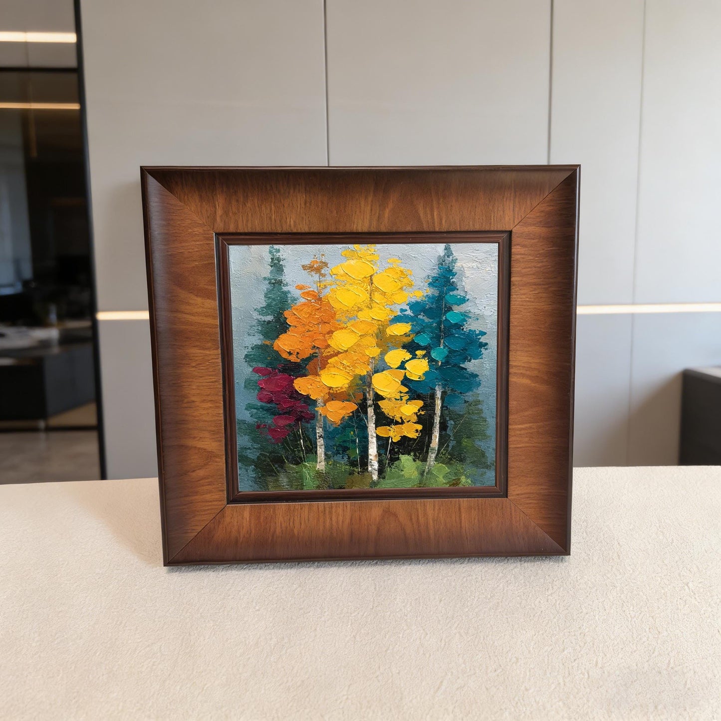 Front-facing view of a square wood-framed autumn landscape print with thick impasto texture, standing on a light-colored surface.