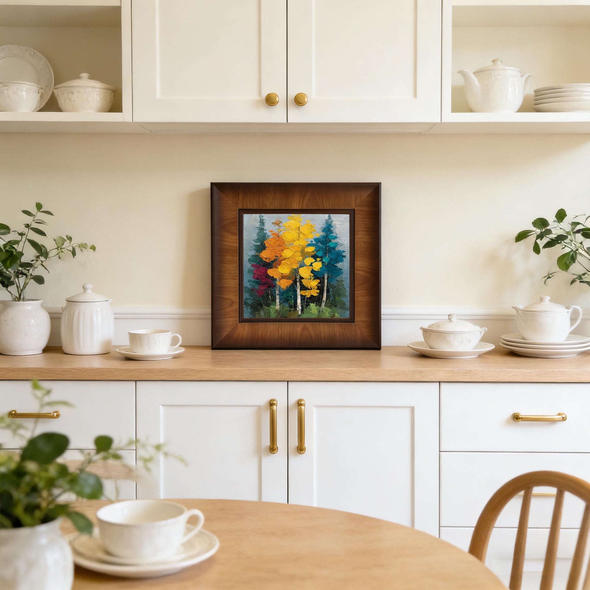 A square wood-framed painting of autumn trees displayed on a wooden countertop in a bright, modern white kitchen.