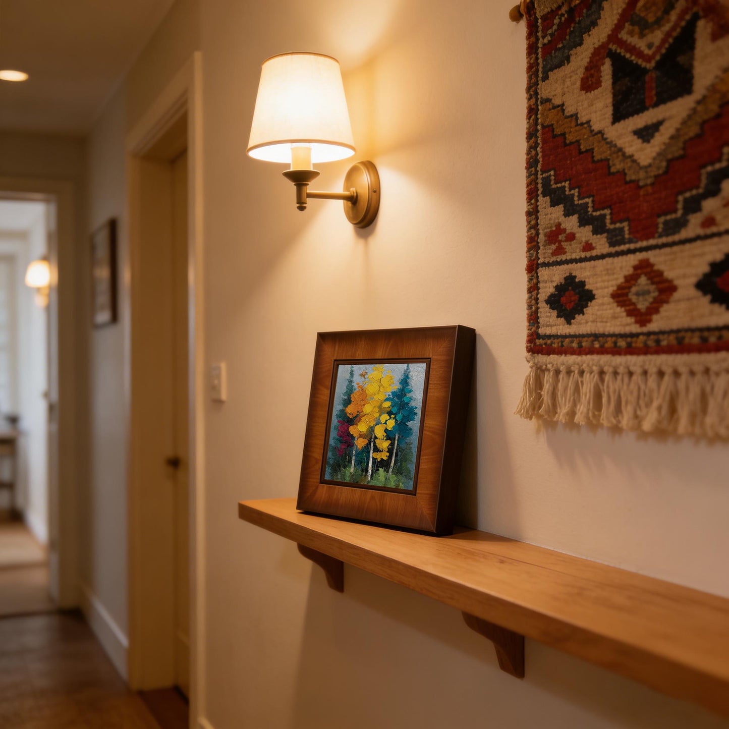 Square wood-framed impasto forest painting sitting on a wooden floating shelf in a hallway, illuminated by a warm wall sconce next to a woven tapestry."