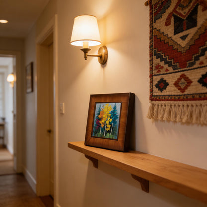 Square wood-framed impasto forest painting sitting on a wooden floating shelf in a hallway, illuminated by a warm wall sconce next to a woven tapestry."