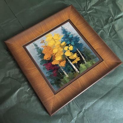 Square wood-framed impasto forest painting sitting on a wooden floating shelf in a hallway, illuminated by a warm wall sconce next to a woven tapestry.