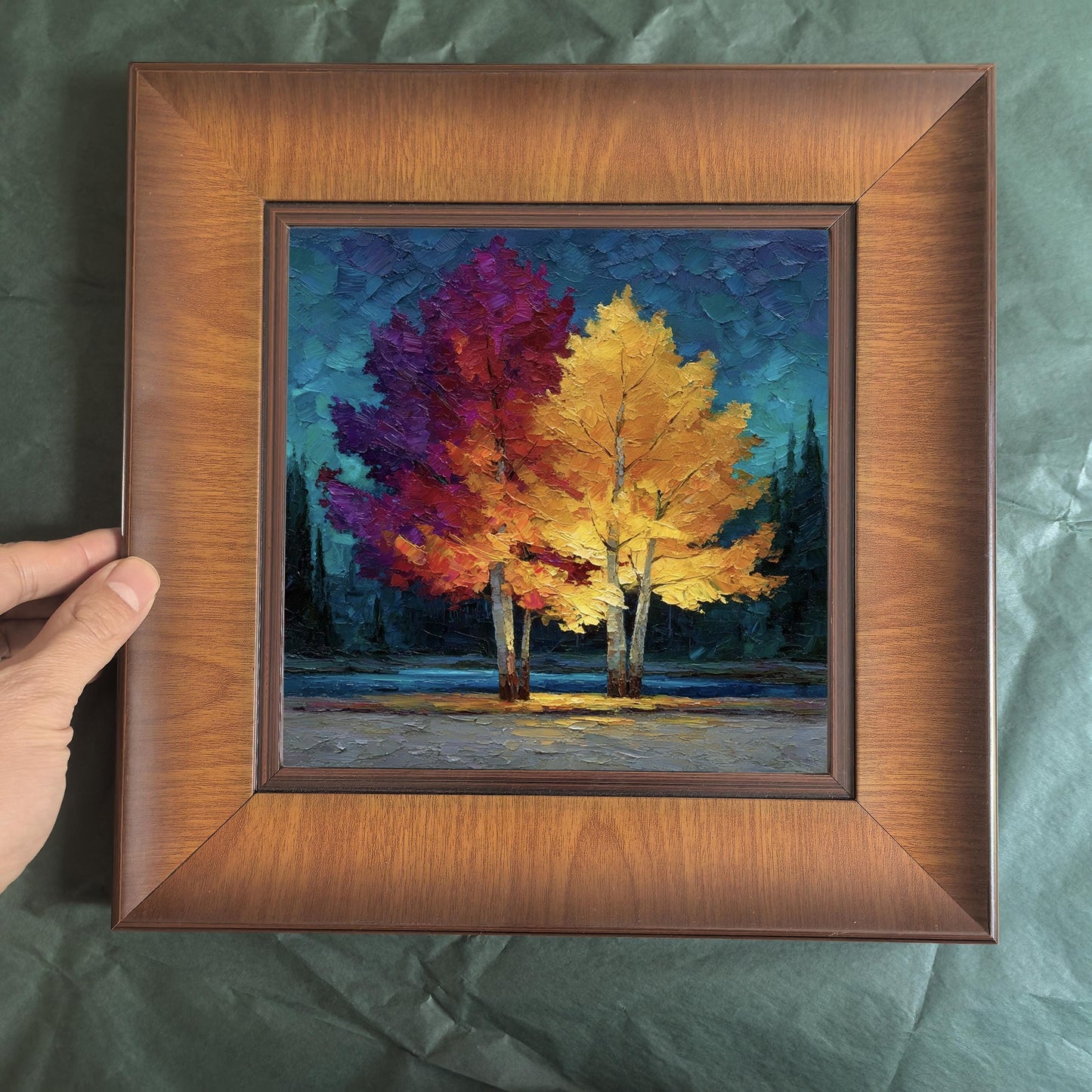 A hand holding the square wood-framed artwork against a dark green textured background, showcasing the frame's depth and sturdy build.