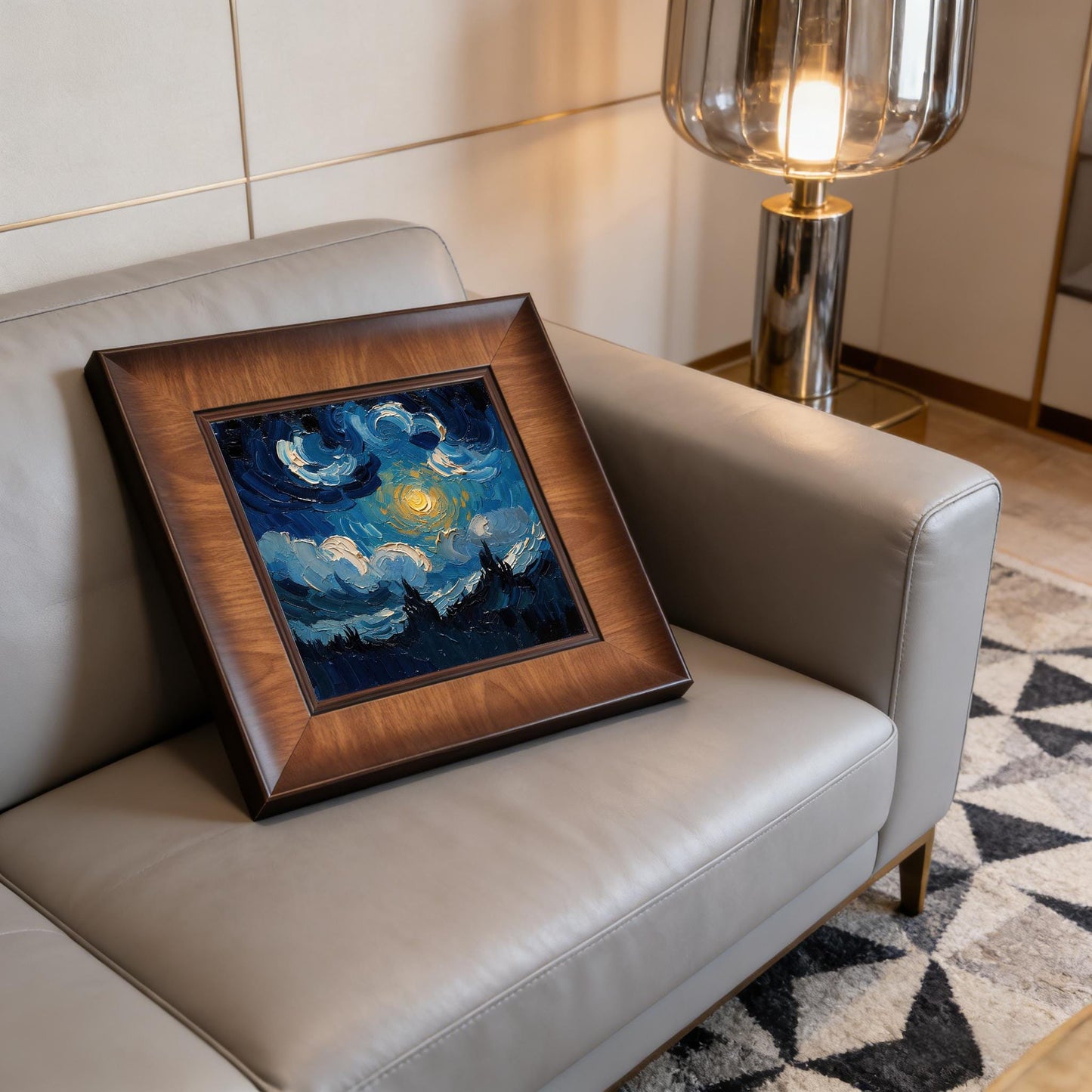 A square wood-framed impasto painting of a starry night sky leaning against the back of a modern grey leather sofa in a well-lit living room with a designer floor lamp.